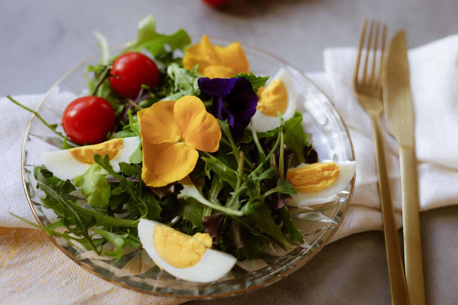 A salad of greens, cherry tomatoes, and boiled egg garnished with yellow and purple pansies on a glass plate