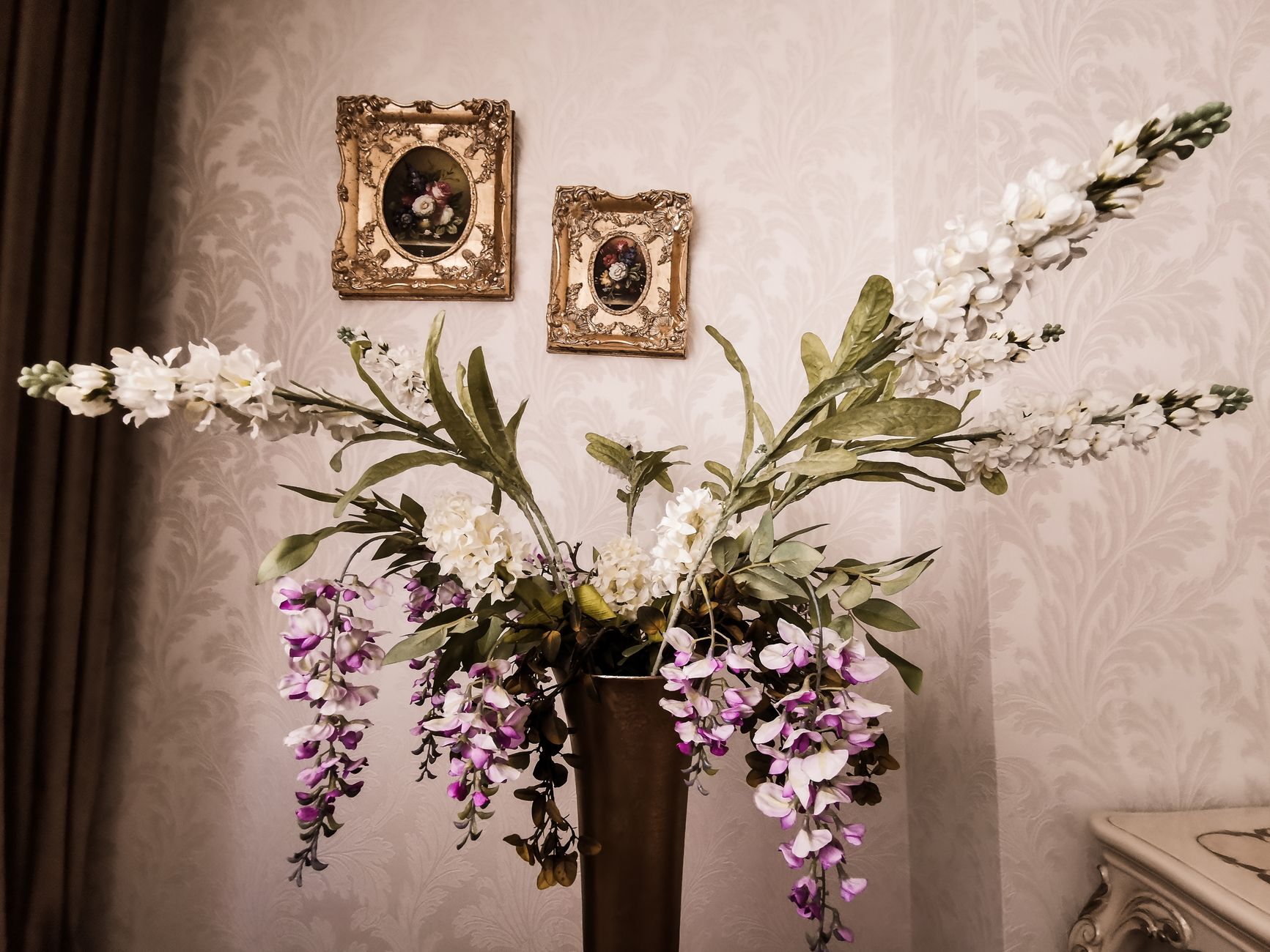 A tall arrangement of white and purple blossoms in a dark vase against a damask wall with small framed paintings