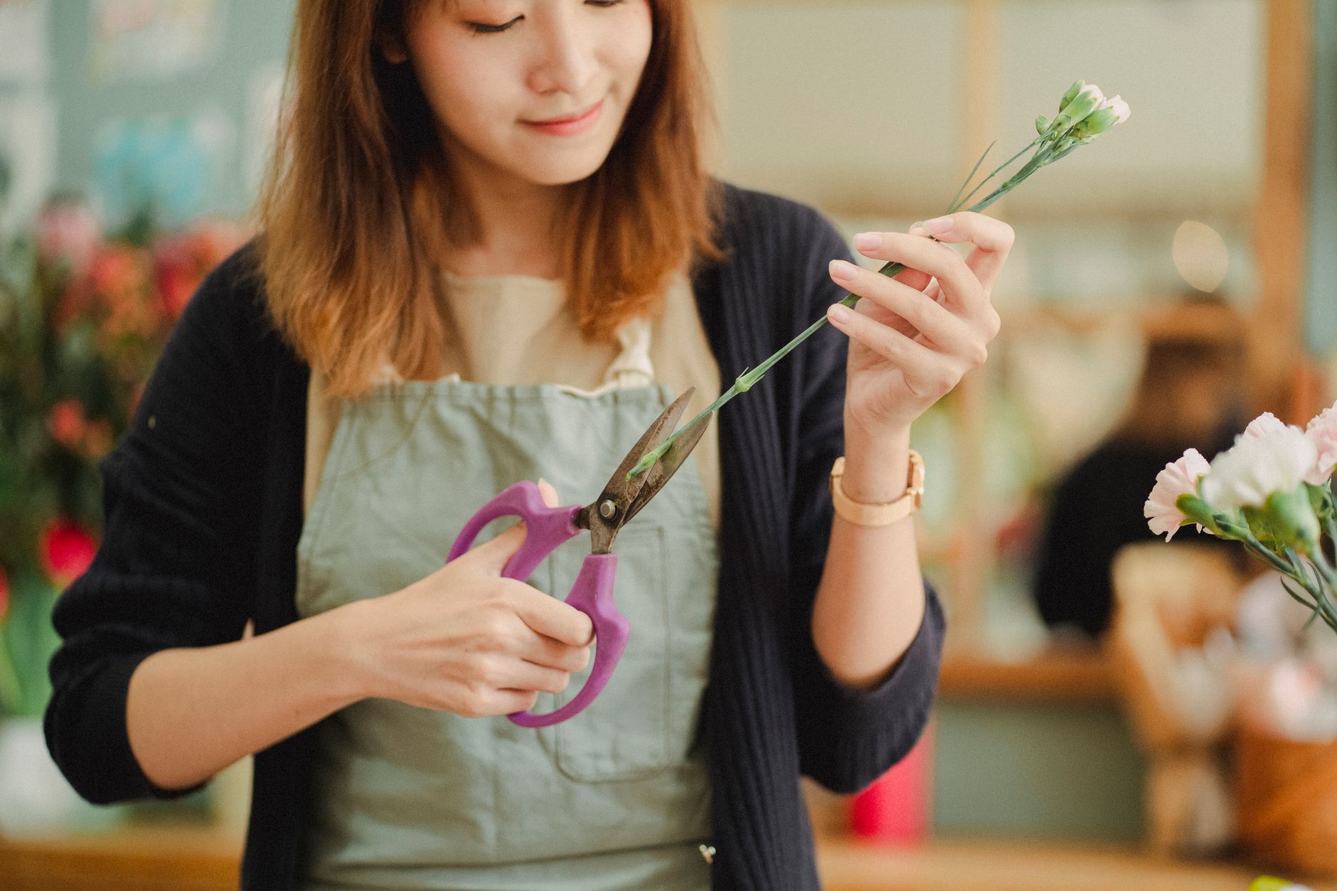 A smiling florist in a linen apron cutting a carnation stem with purple-handled scissors
