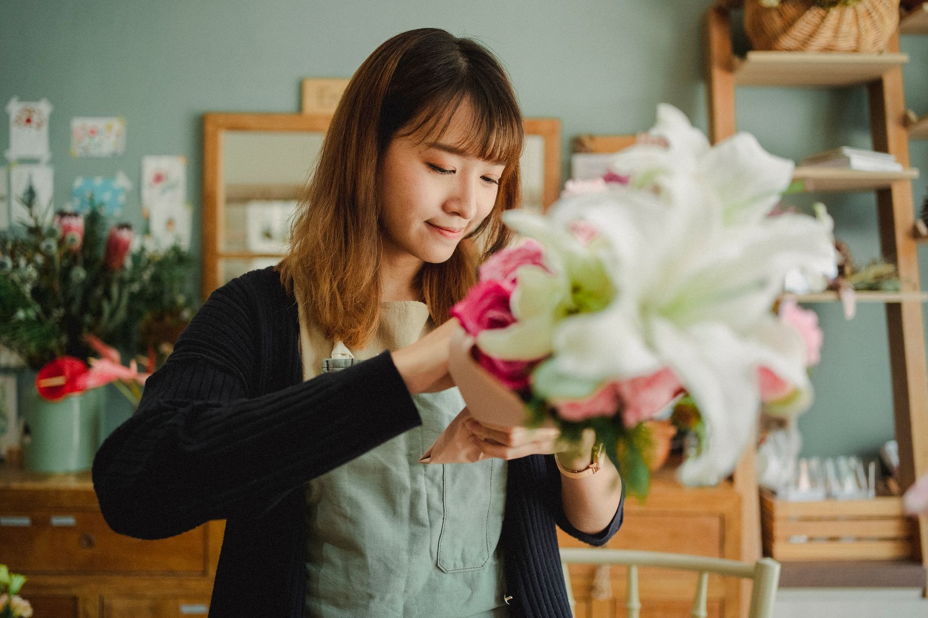 A florist in an apron smiling as she arranges a bouquet of white lilies and pink flowers in her shop