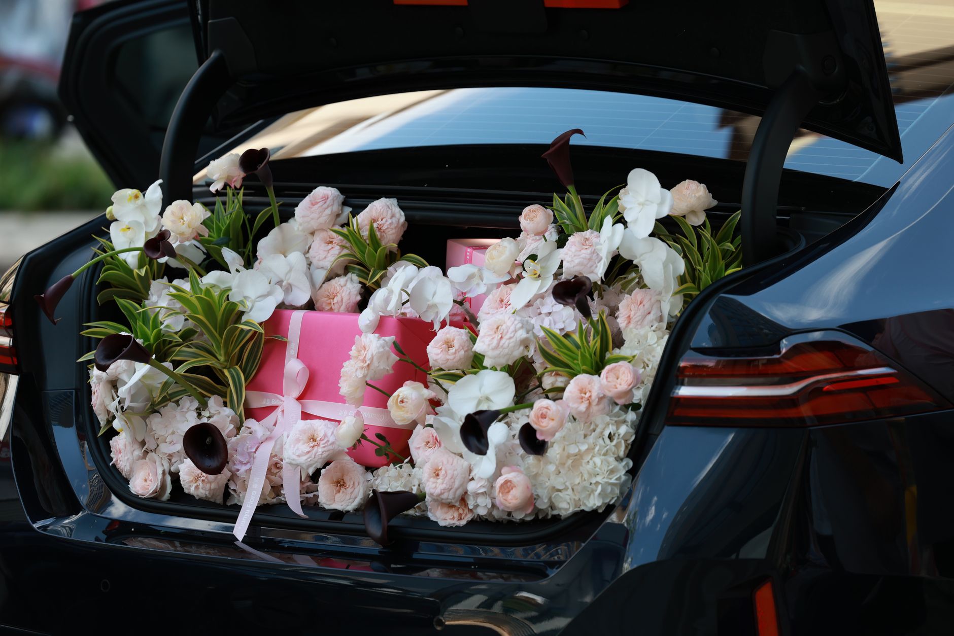 A large floral arrangement of white and blush flowers and a pink gift box carefully packed in an open car trunk