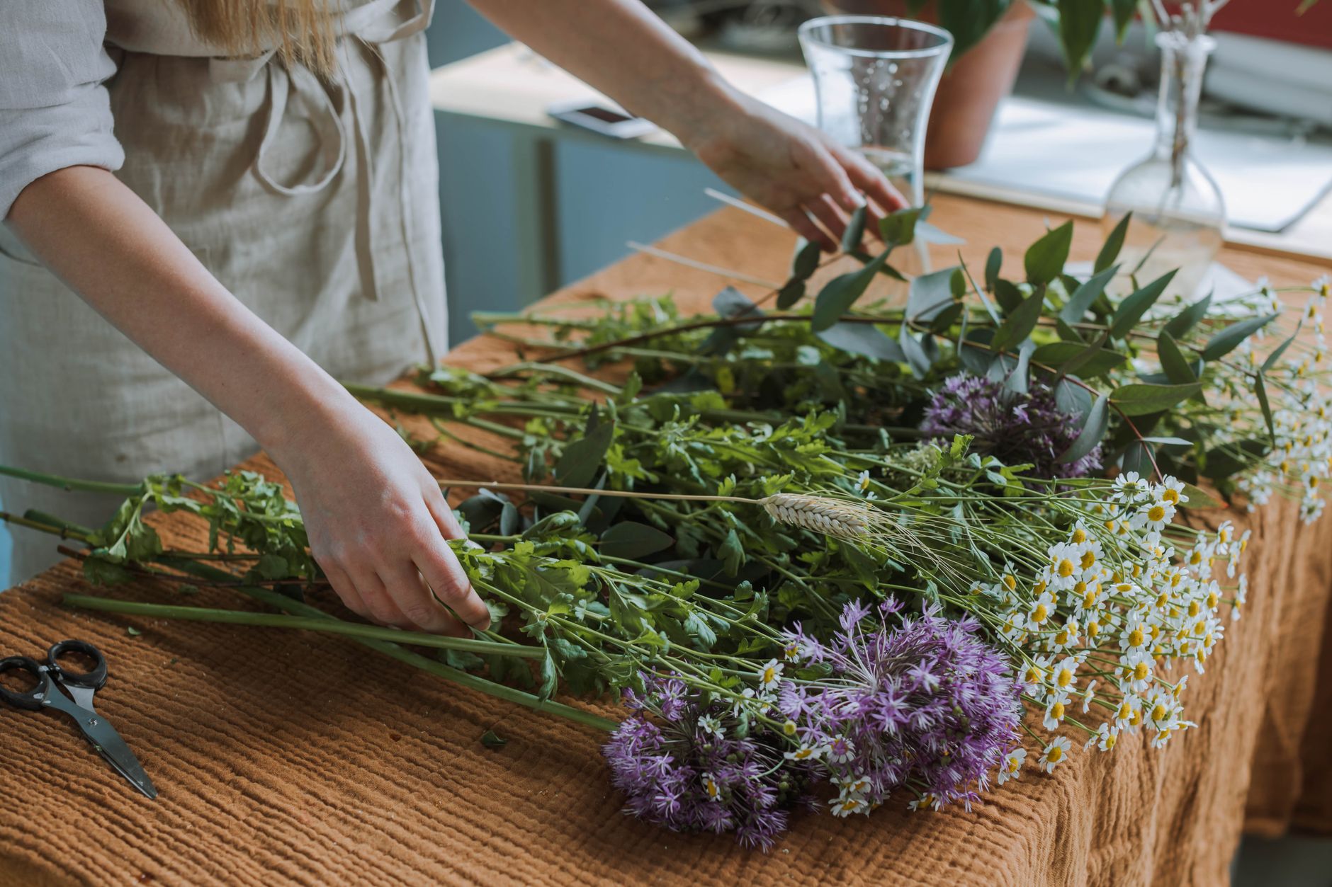 A florist in a linen apron arranging daisies, alliums, and greenery on a table, with scissors nearby