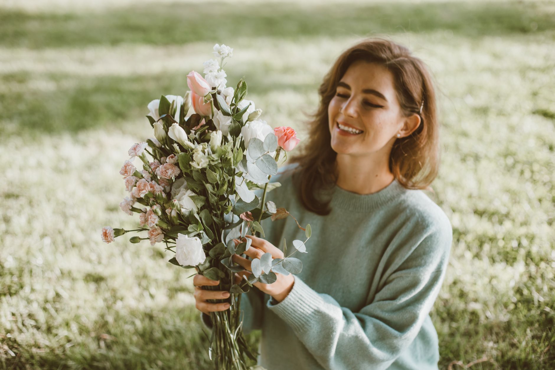 A smiling woman in a sage sweater holding a loose bouquet of pink and white flowers with eucalyptus in a sunny park