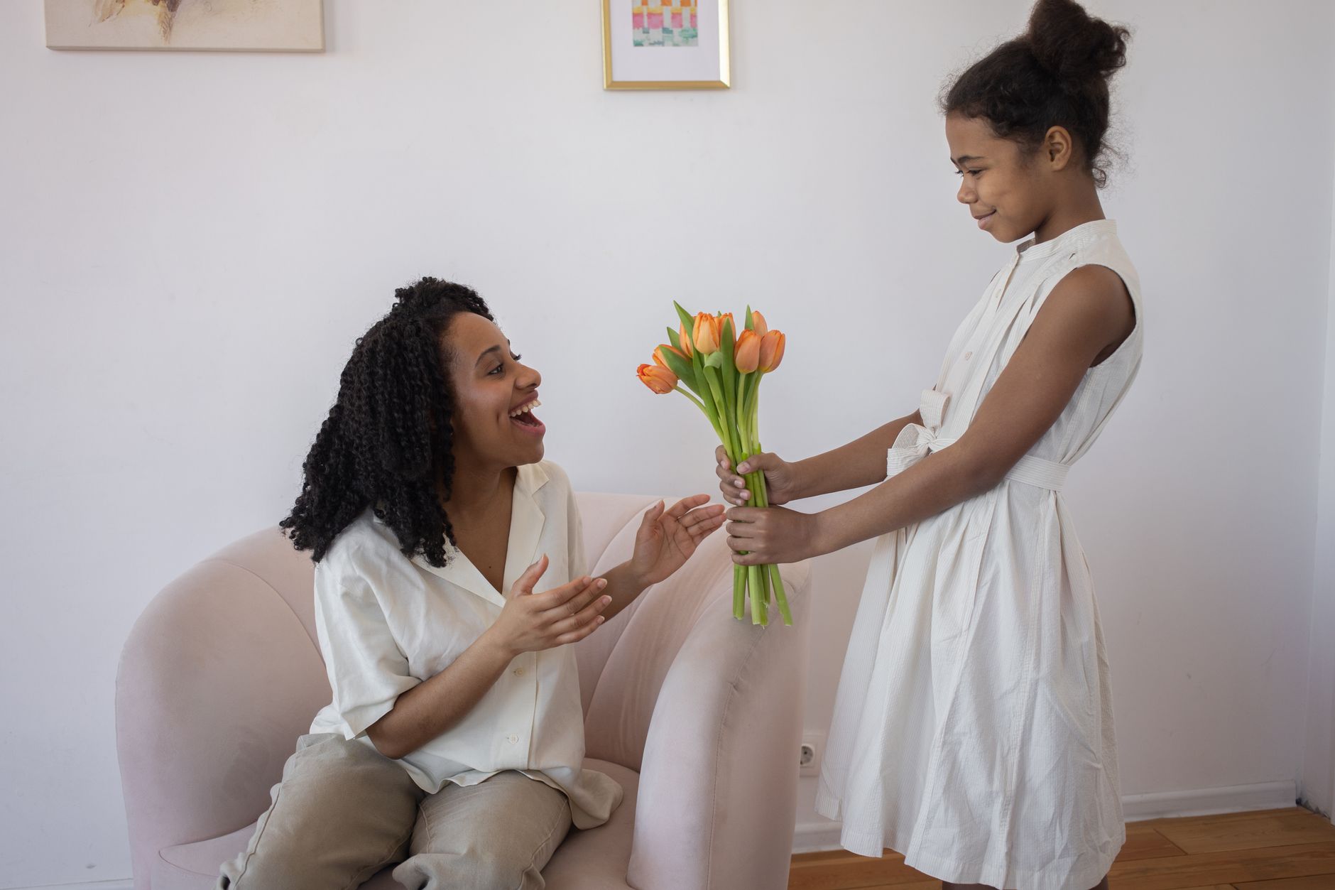 A girl in a white dress hands a bouquet of orange tulips to a delighted woman seated in an armchair