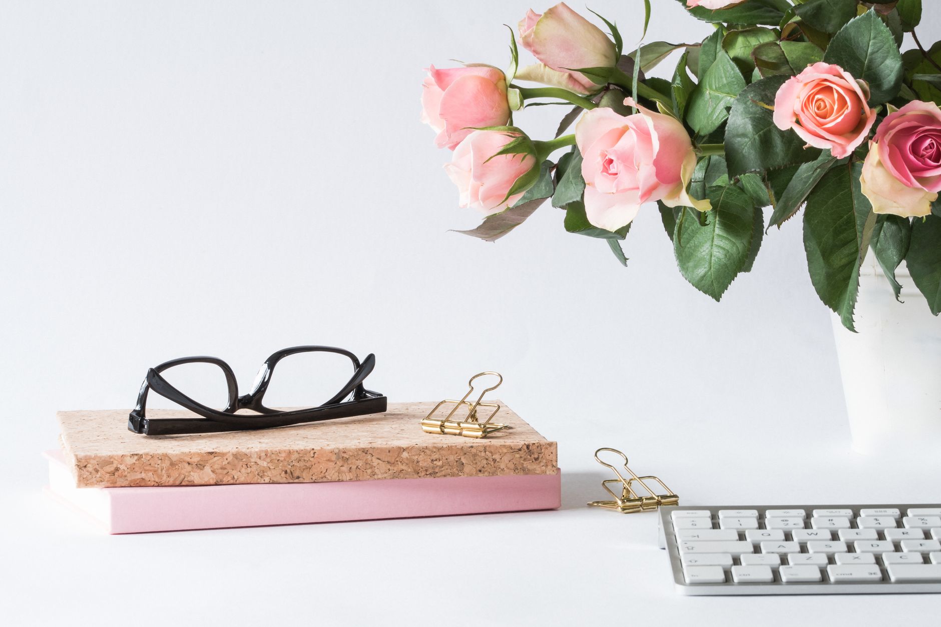 Pink roses in a white vase beside black glasses, a notebook, and a keyboard on a bright desk