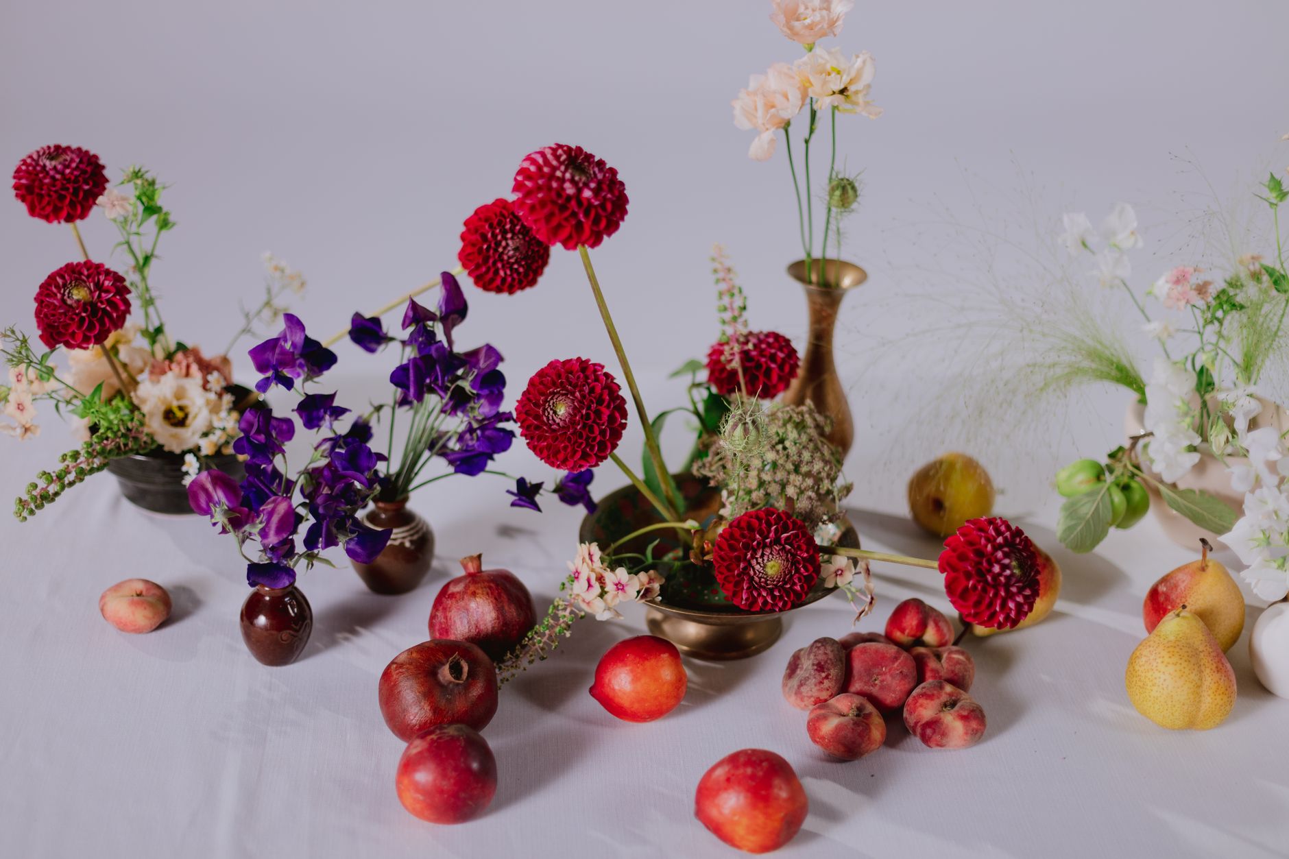 A table with dahlias, sweet peas, and other cut flowers arranged among pomegranates, peaches, and pears