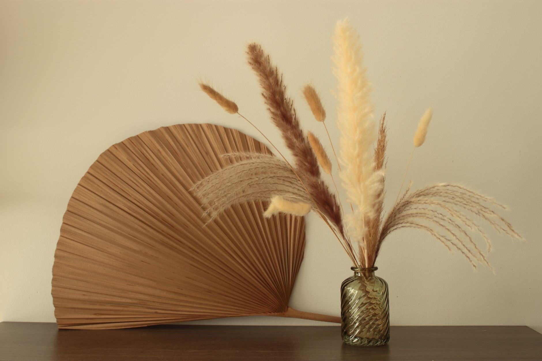 Dried pampas grass and bunny-tail grass in a small green glass vase beside a woven palm fan