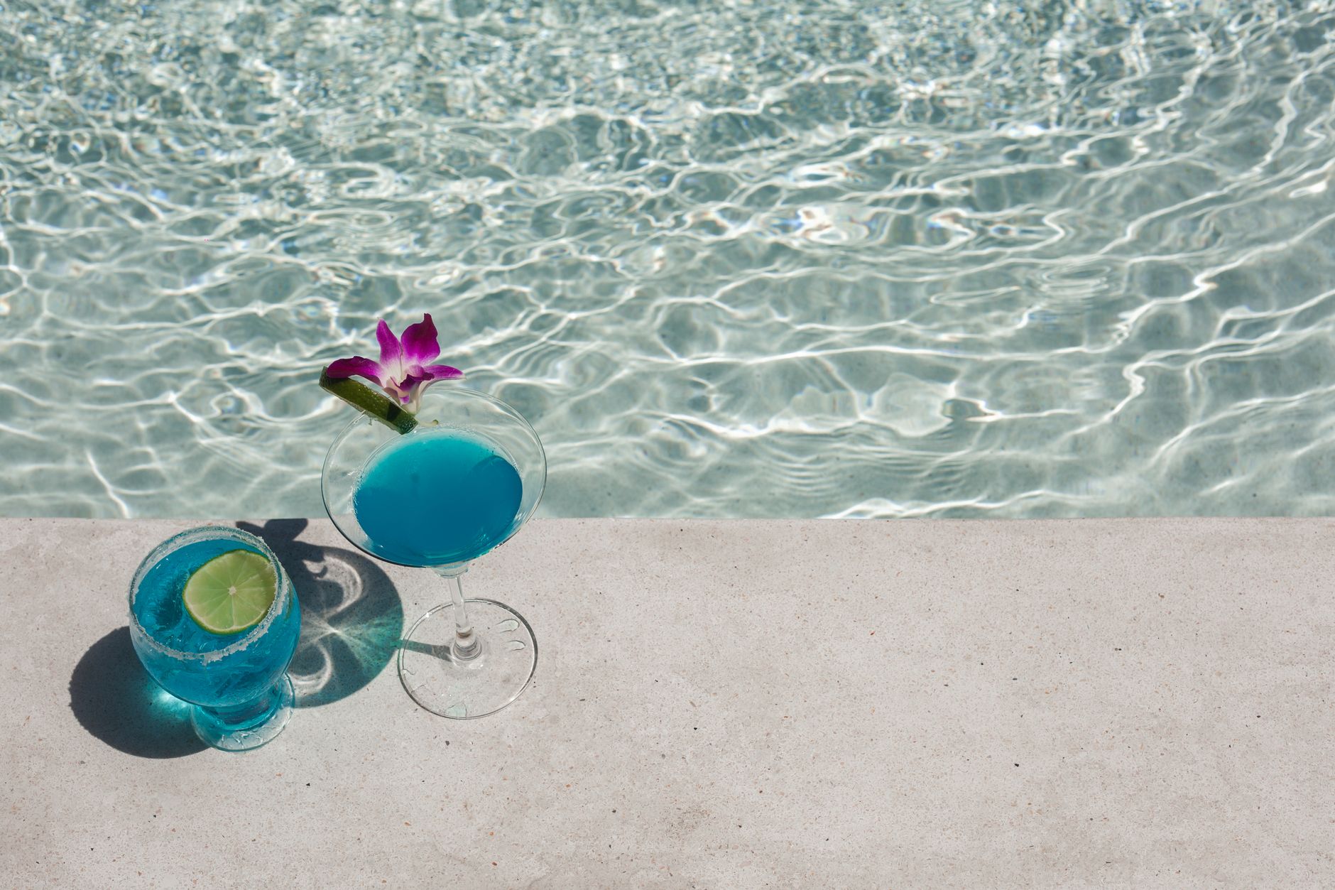 Two blue cocktails, one with a lime wheel and one with a small purple orchid, by a sunlit pool