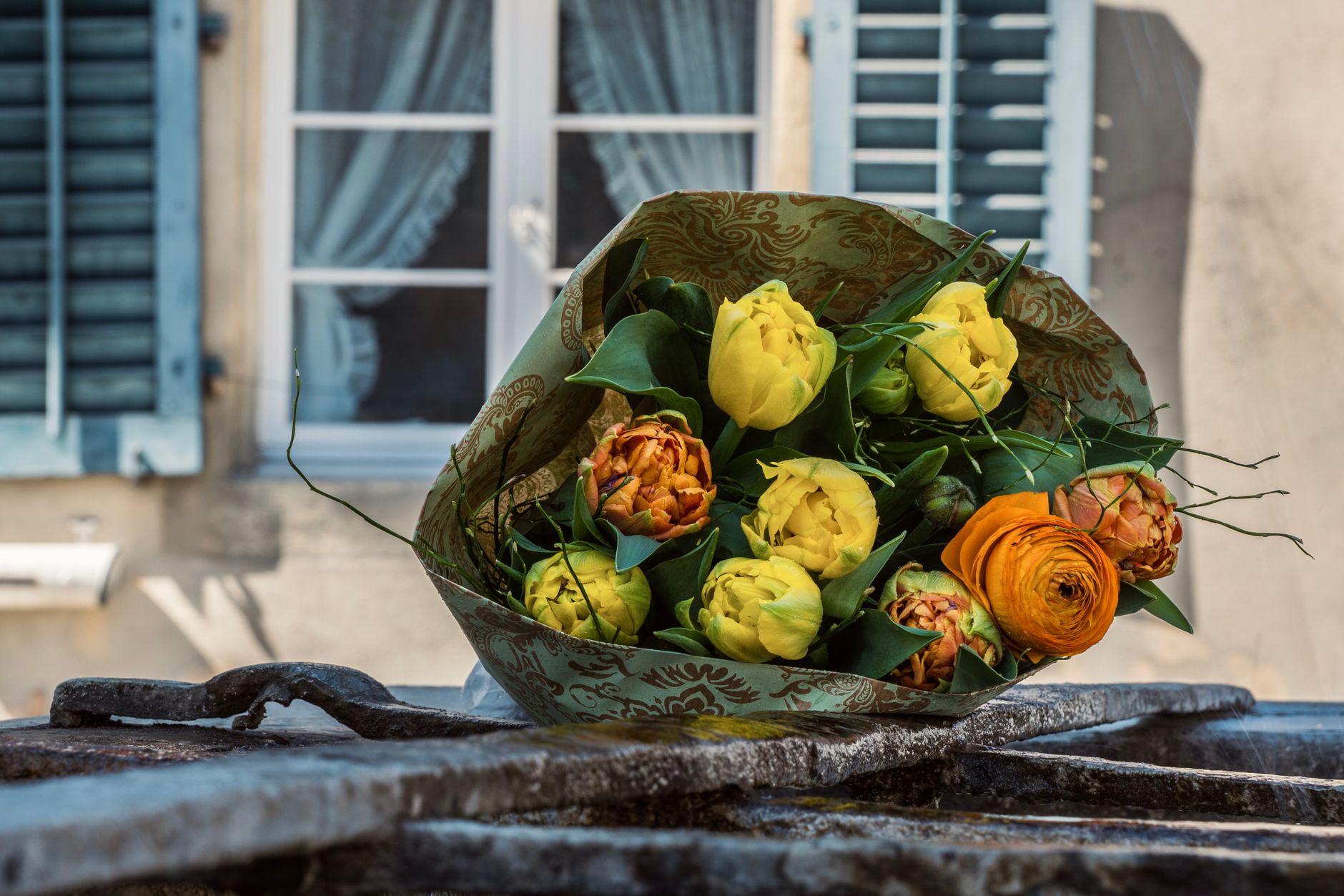A wrapped bouquet of yellow and orange tulips resting on a stone ledge in front of a shuttered window