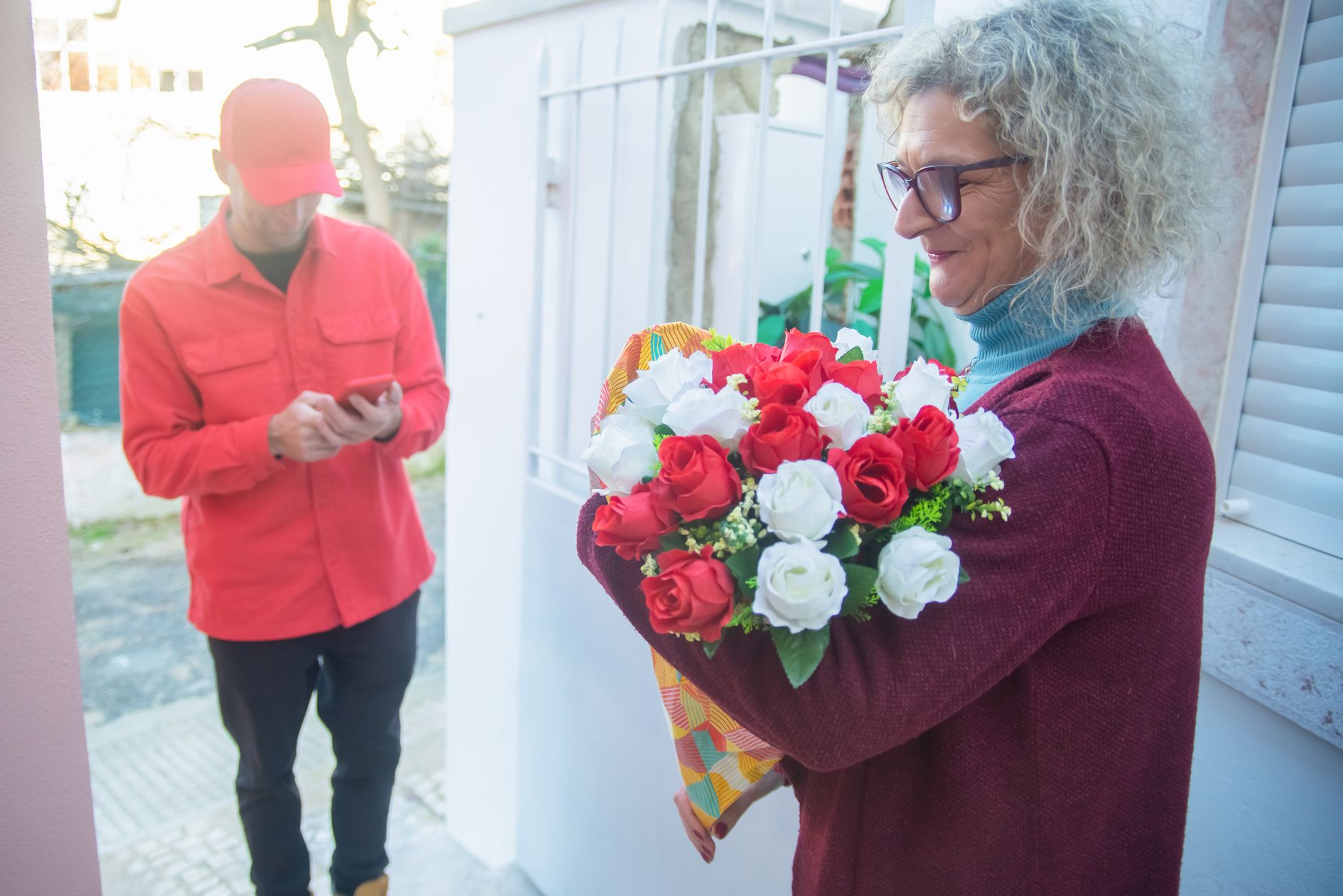 A smiling woman in glasses receives a bouquet of red and white flowers from a courier at her door