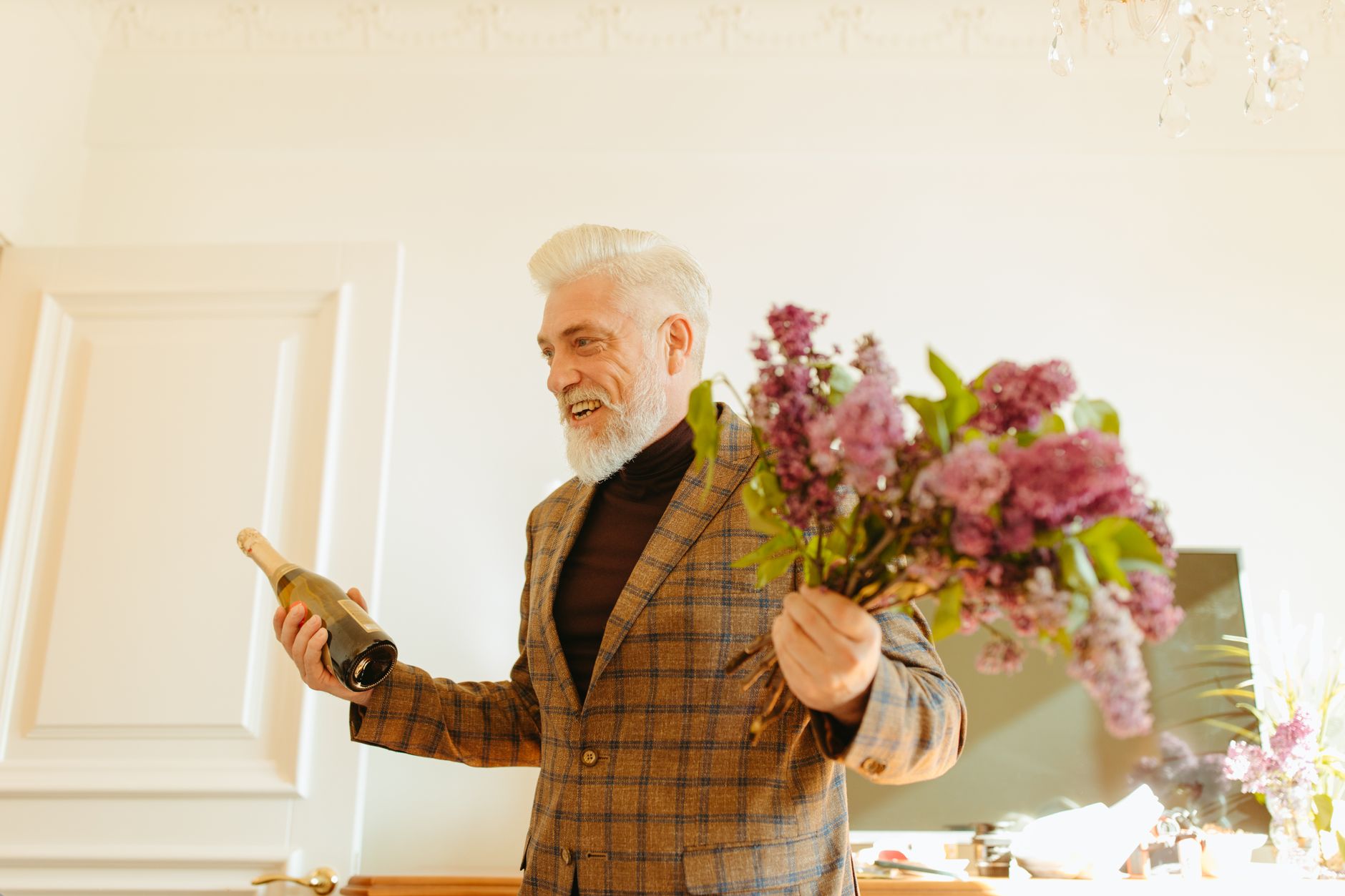 A smiling gray-bearded man in a plaid blazer holding a bunch of lilac flowers and a bottle of sparkling wine