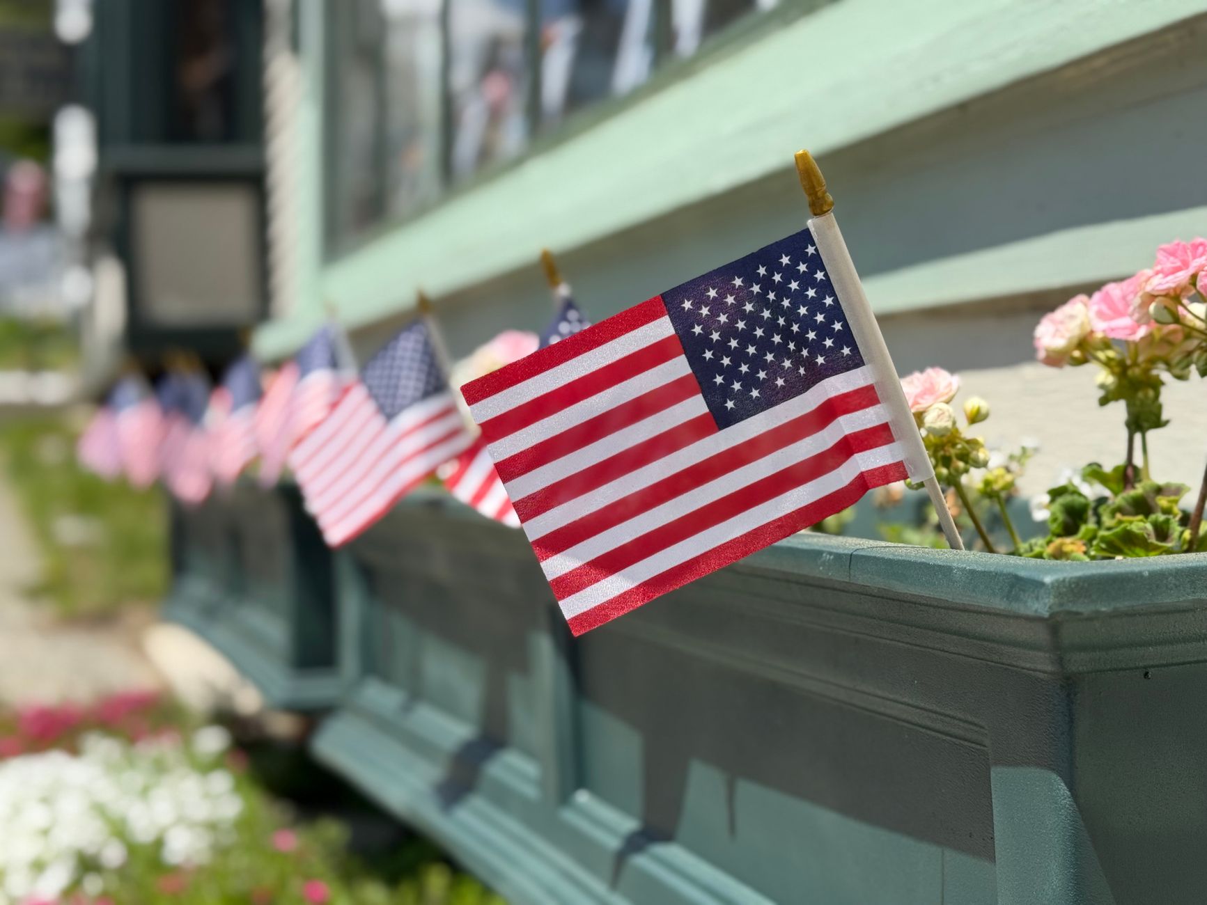 A row of small American flags in a teal window box with pink geraniums