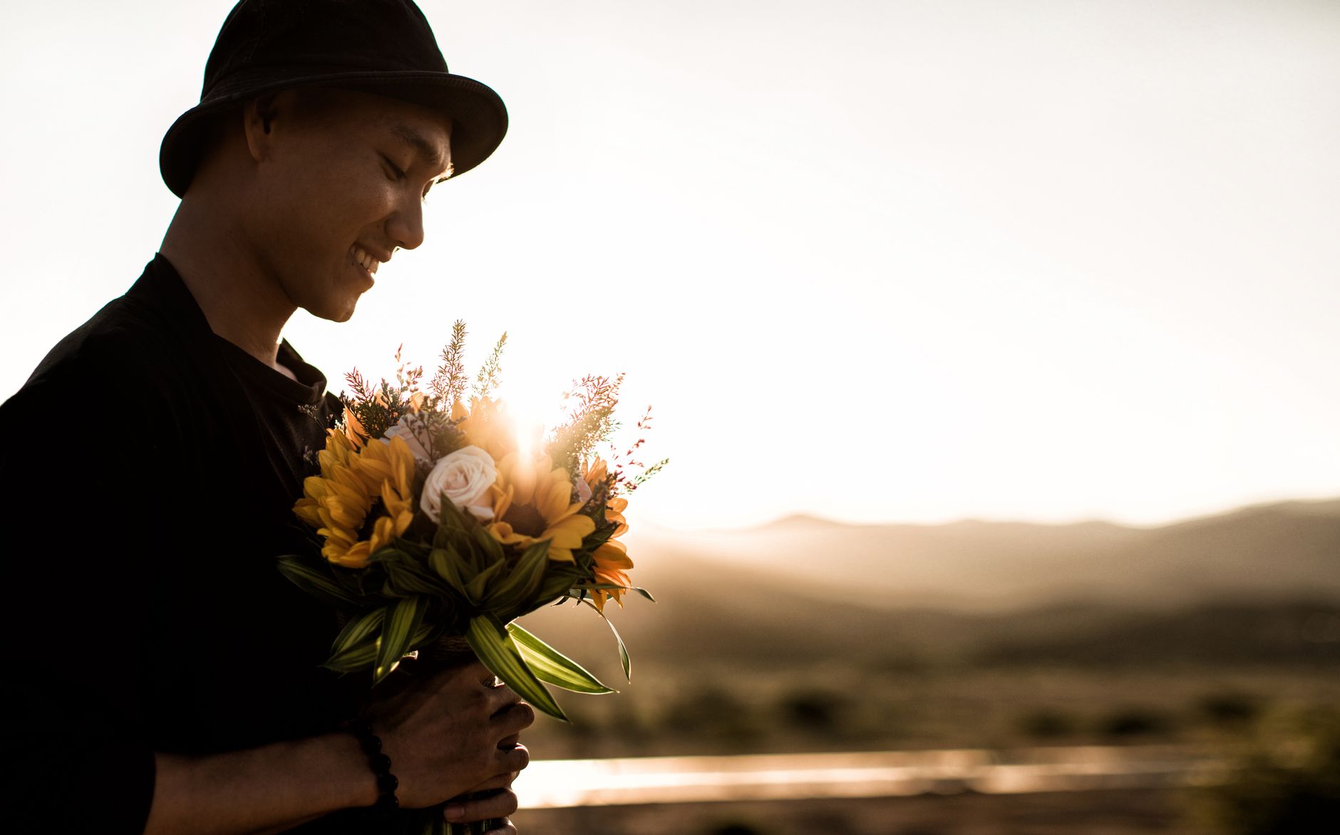 A smiling man in a brimmed hat holding a bouquet of sunflowers and a cream rose at sunset