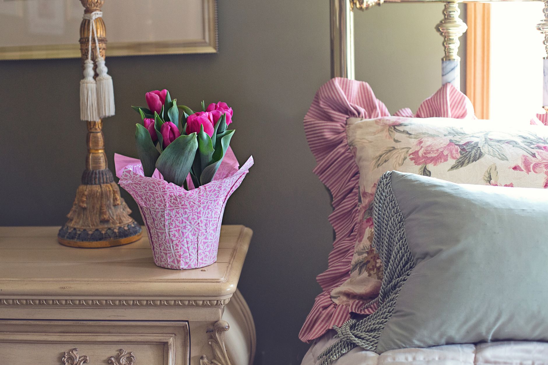 A small vase of white blossoms on a bedside table next to a bed with white linens and a sunlit curtain