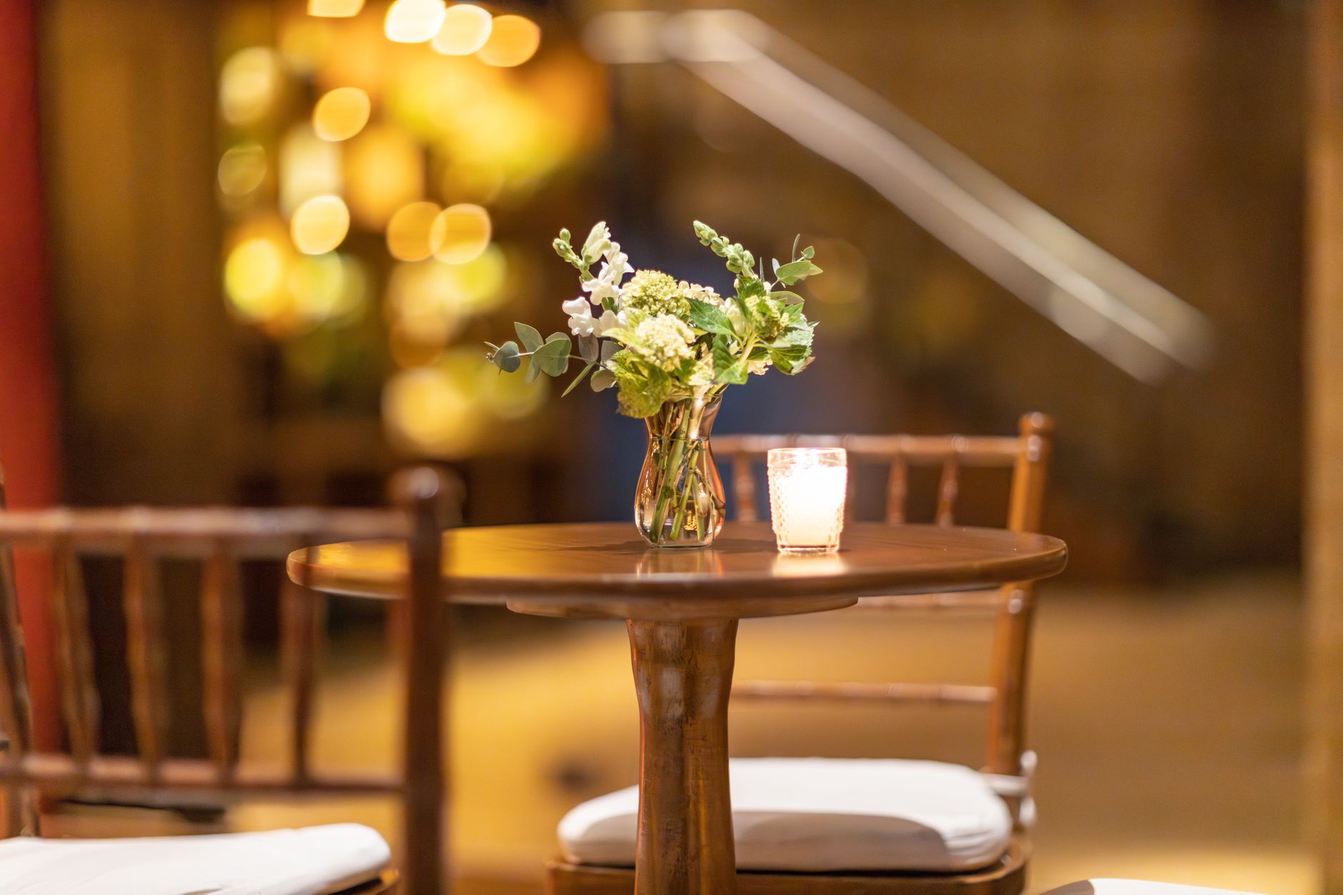 A small glass vase of white and green flowers beside a lit candle on a round wooden table in a warmly lit restaurant