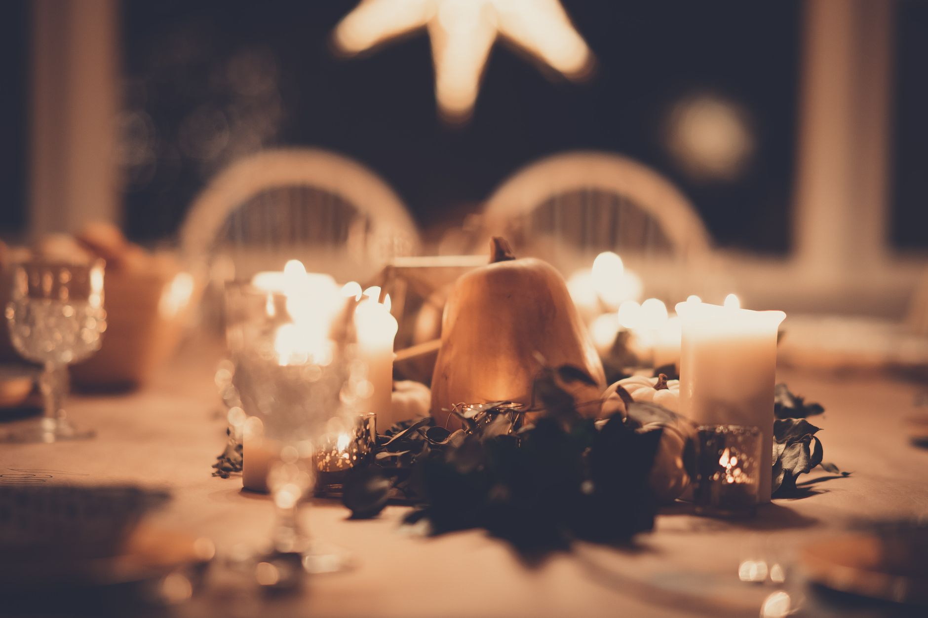 A candlelit Thanksgiving table with a low pumpkin and greenery centerpiece, glowing candles, and crystal glasses