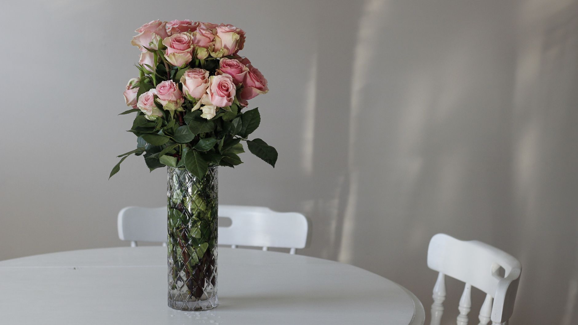 A glass vase of soft pink roses on a white table in a quiet, sunlit room