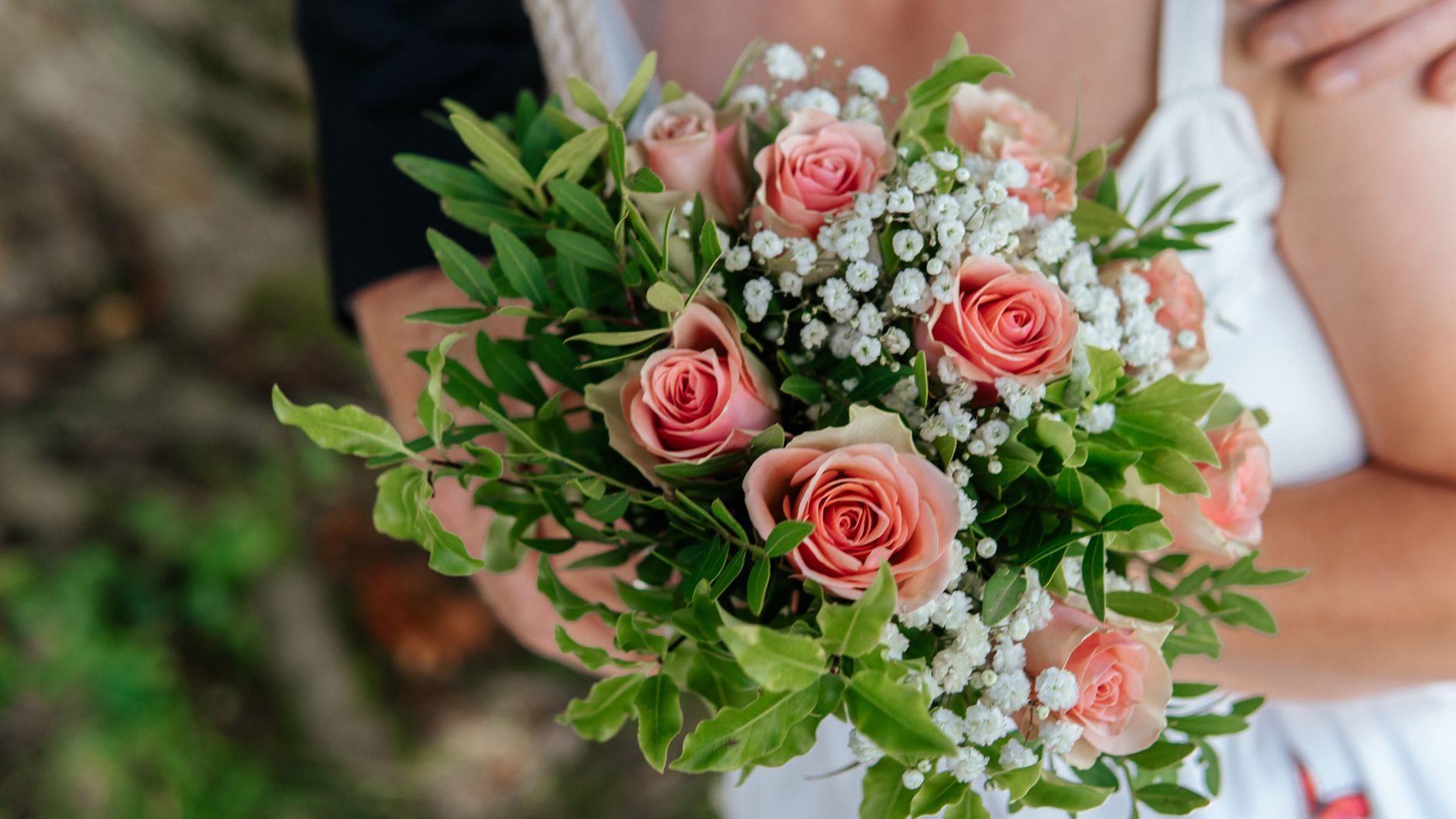 A bridal bouquet of pink roses, baby's breath, and green leaves held against a white dress