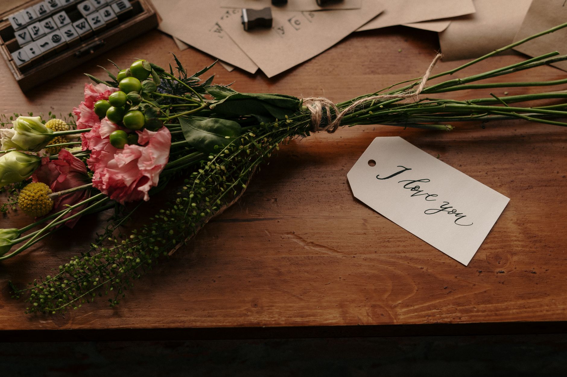 A hand-tied bouquet of pink and green flowers tied with twine on a wooden table, beside a handwritten I love you tag