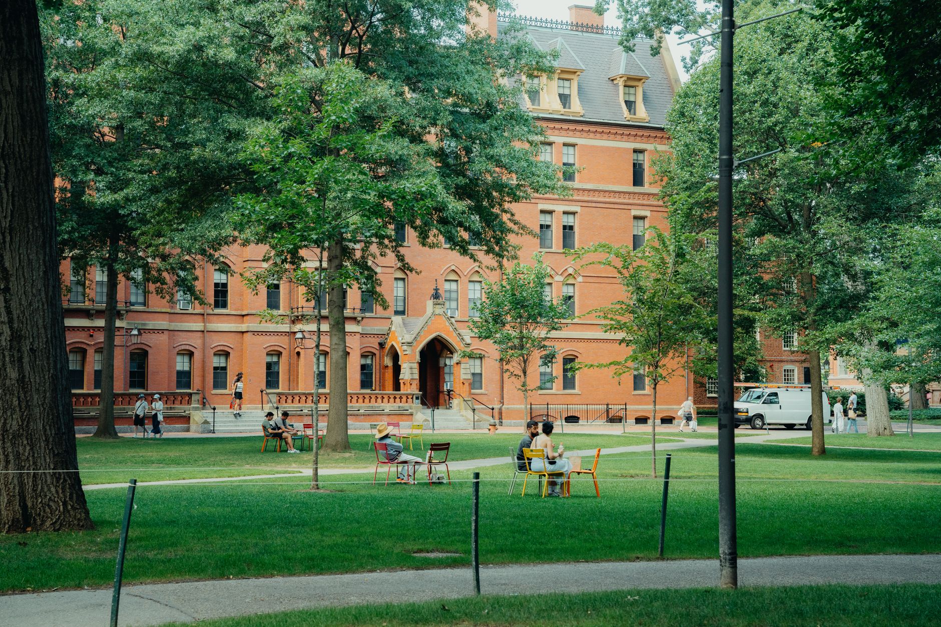 Students sitting on chairs on a lawn in front of a red brick university building with large shade trees