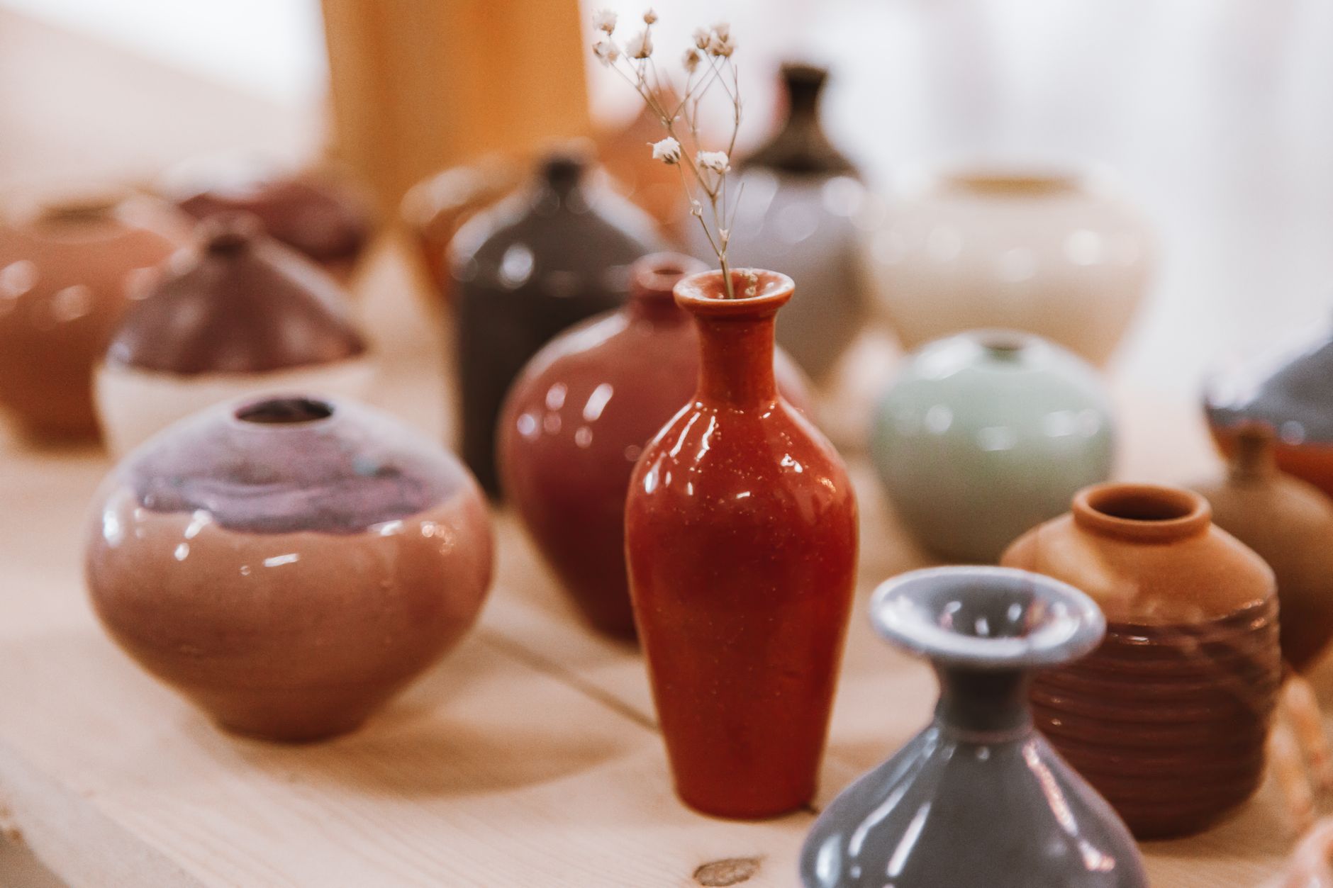 A table full of glossy handmade ceramic vases in red, gray, brown, and green with dried stems in one
