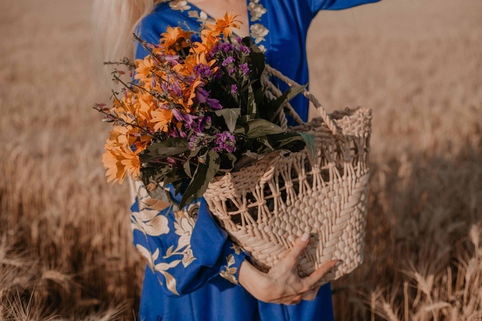 A woman in a blue floral dress holding a woven basket full of orange and purple wildflowers in a golden field