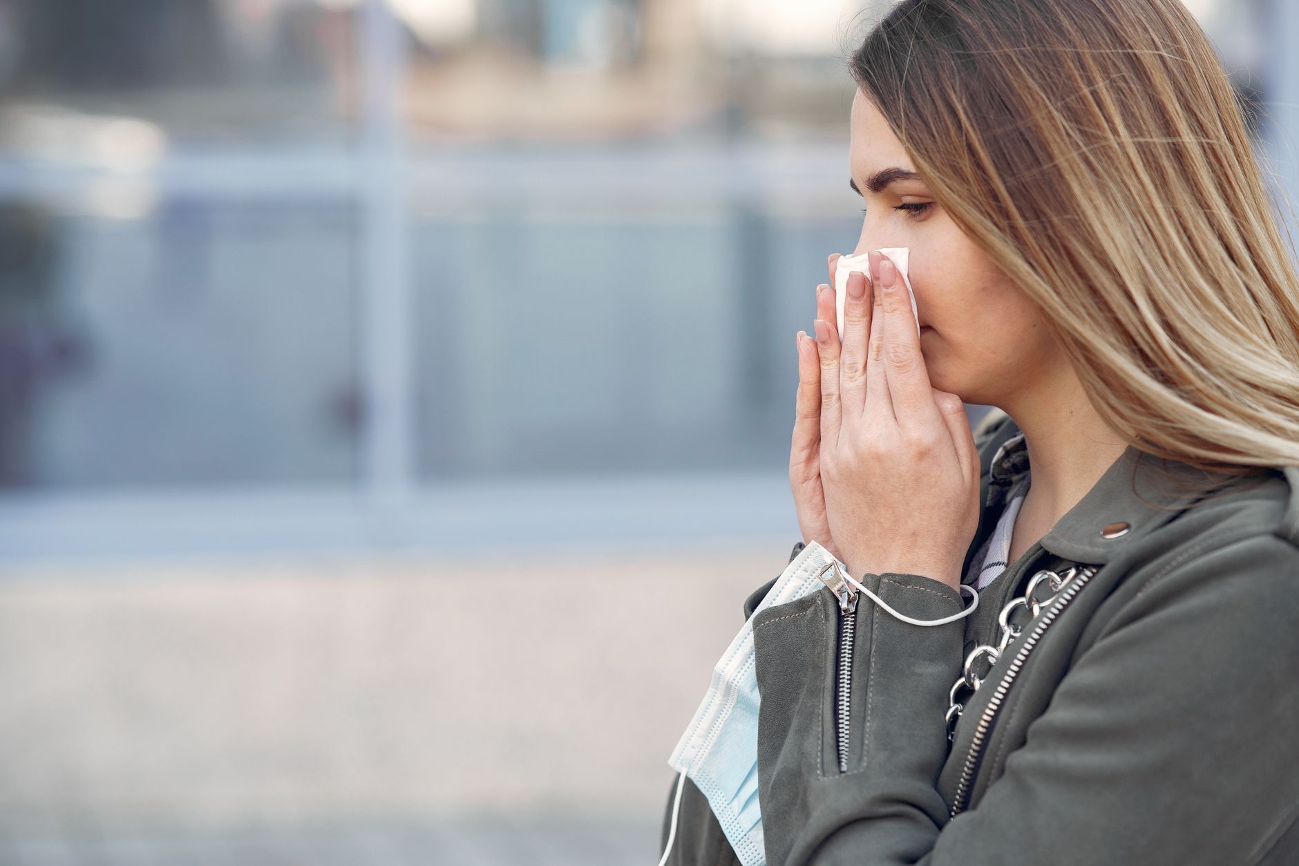 A woman outdoors holding a tissue to her nose with a face mask around her wrist