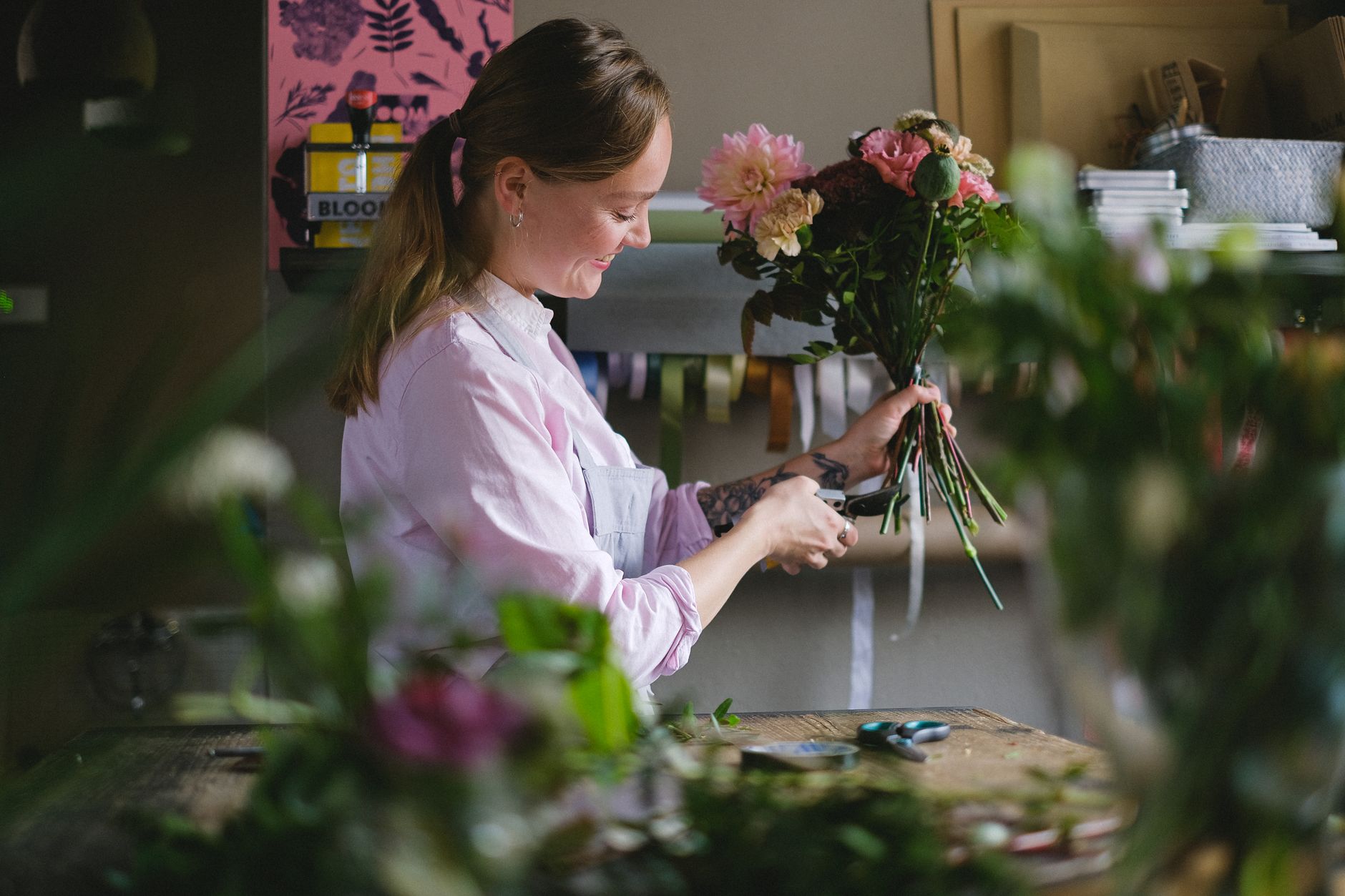 A florist in a pink shirt arranging a bouquet of pink dahlias and roses at a workbench