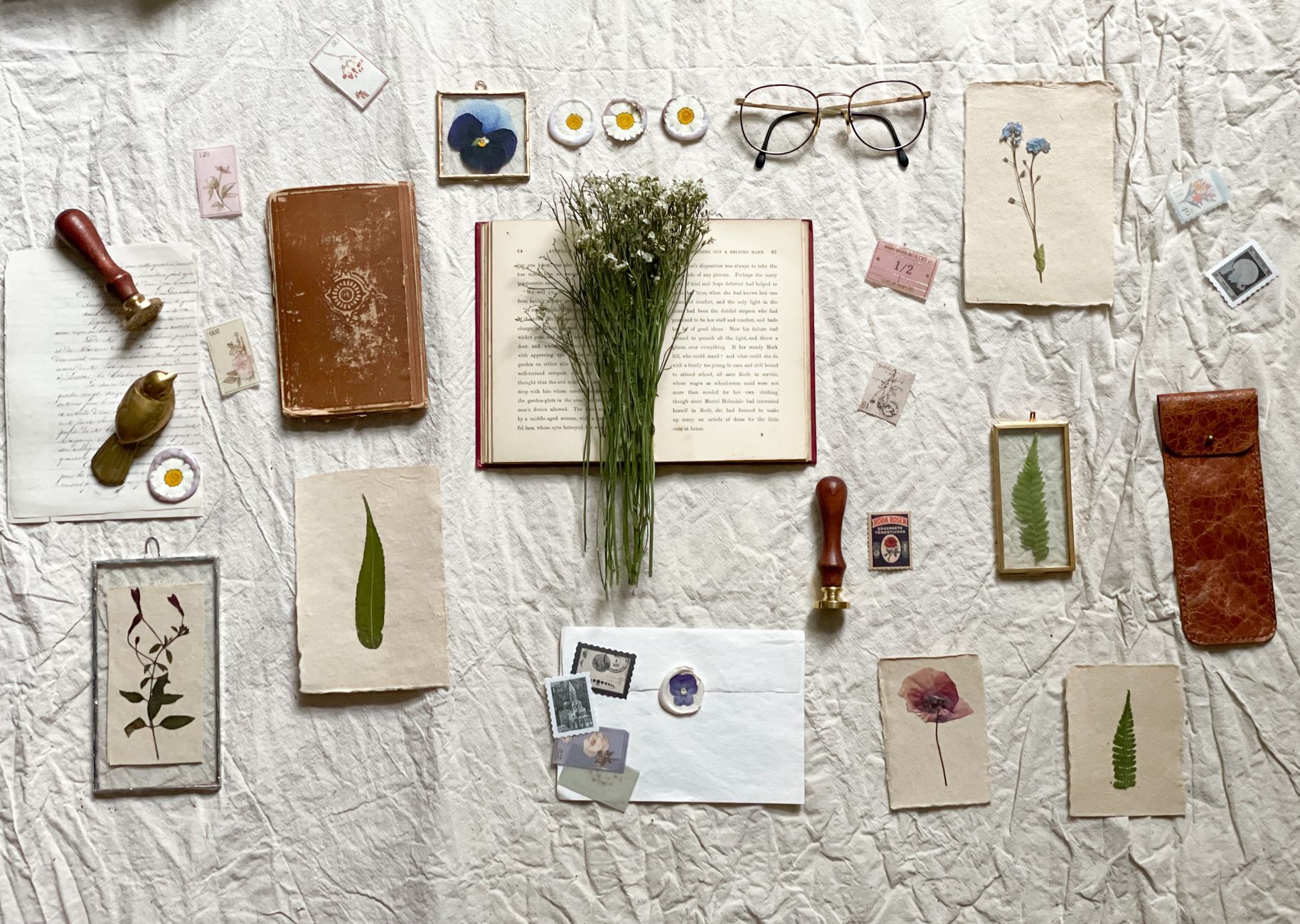 A flat lay of dried and pressed flowers, an open book, antique stamps, and a pair of round glasses on linen
