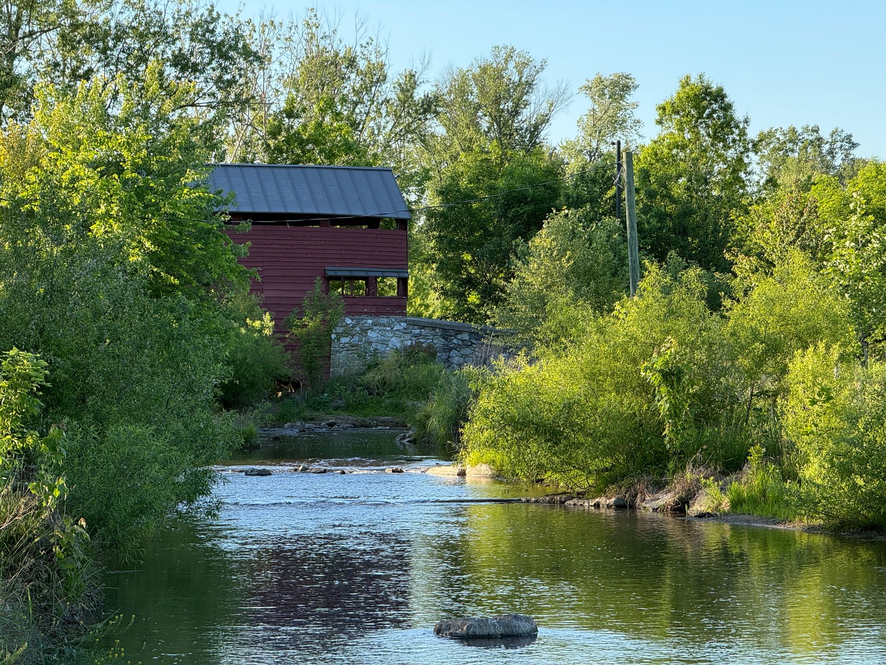 A red covered bridge beside a calm stream surrounded by green trees in summer