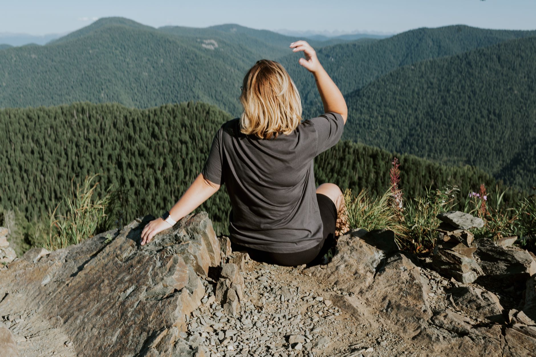 A hiker with blond hair sitting on a rocky summit above a forested mountain valley