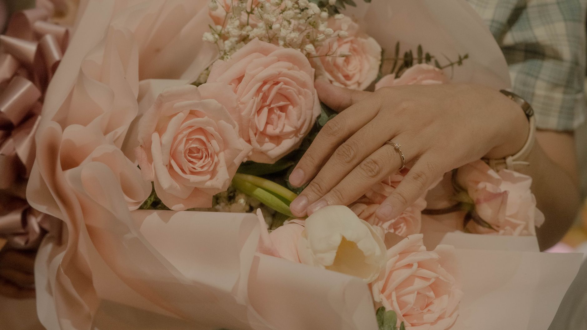 A hand wearing an engagement ring resting on a bouquet of pale pink roses wrapped in pink paper