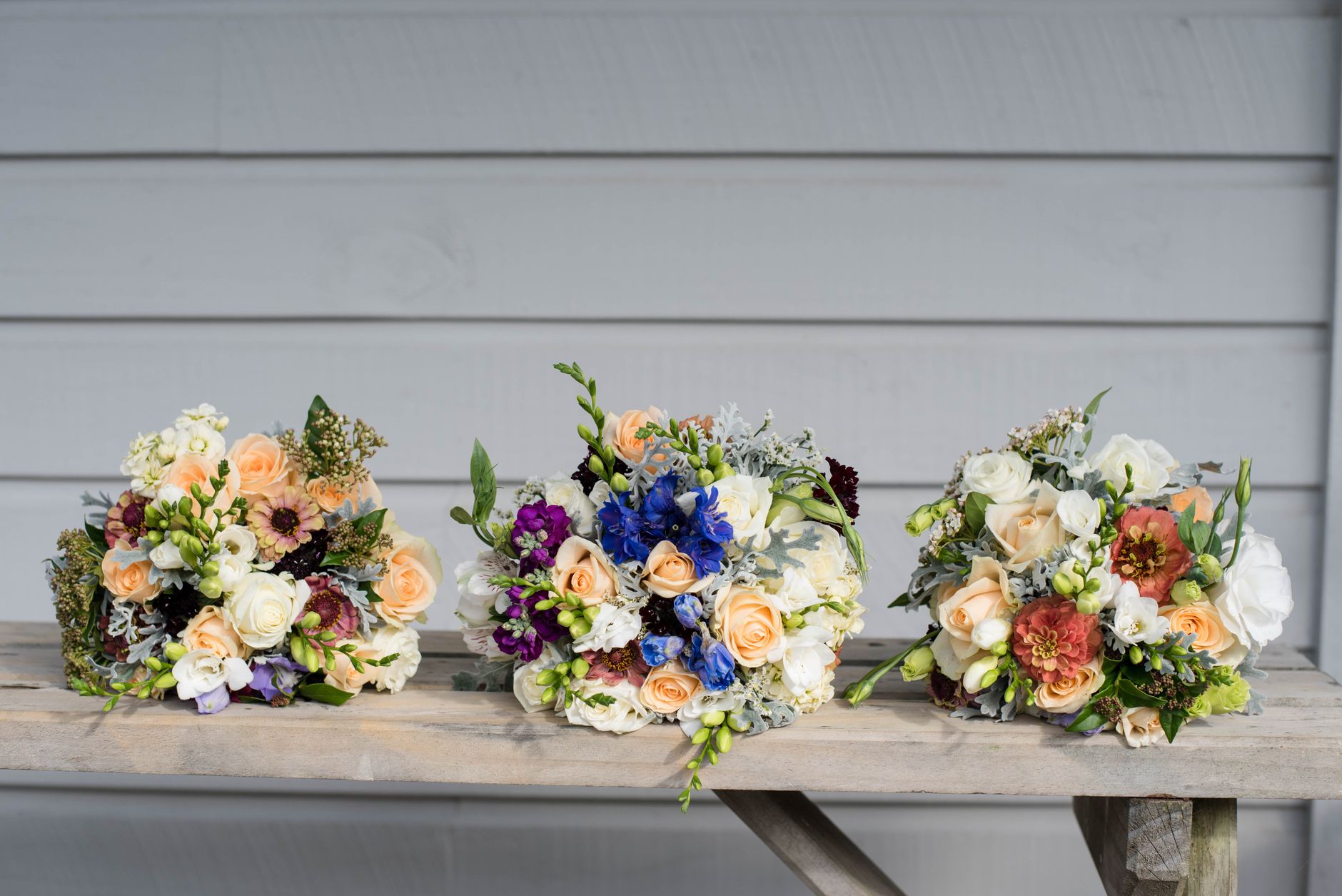 Three bouquets of cream, orange, and purple flowers lined up on a wooden bench in front of gray siding
