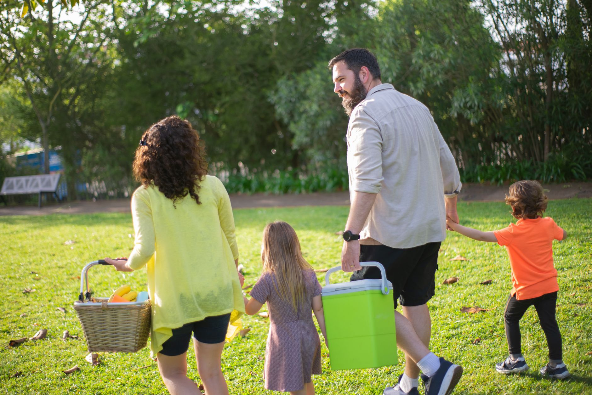 A family of four carrying a picnic basket and a green cooler across a sunny park lawn