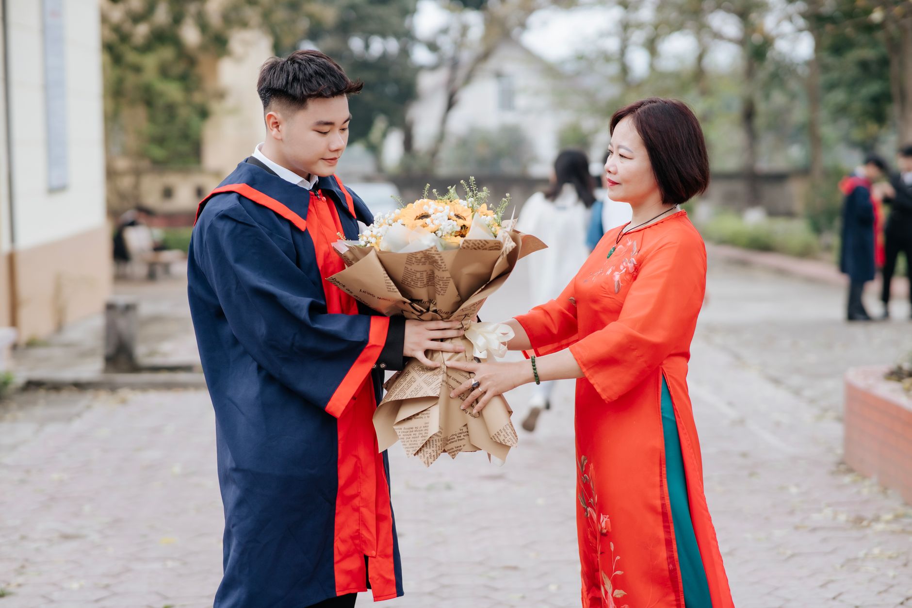 A young graduate in a navy gown receiving a wrapped bouquet from an older woman in an orange dress