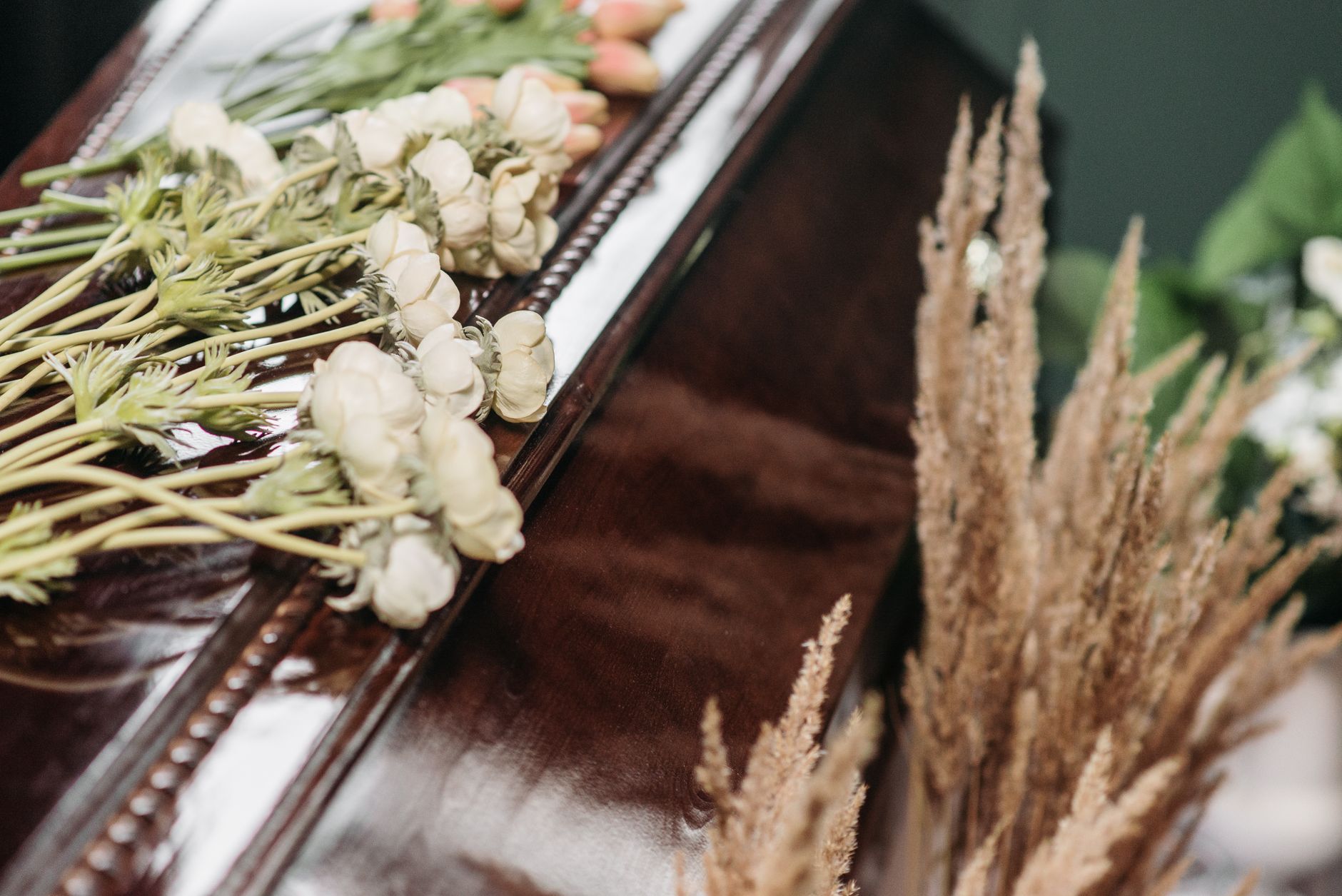 White roses and baby&rsquo;s breath resting on a polished wooden casket beside dried grasses