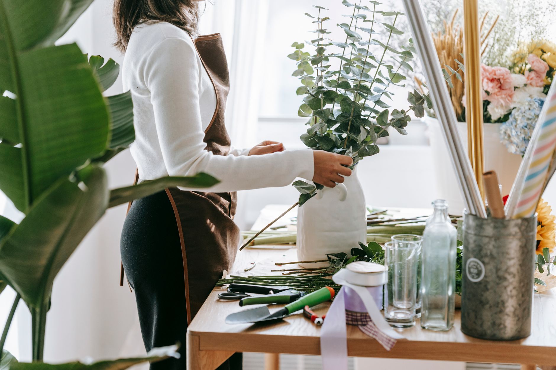A florist in an apron arranging eucalyptus in a white vase at a workbench with tools and ribbons