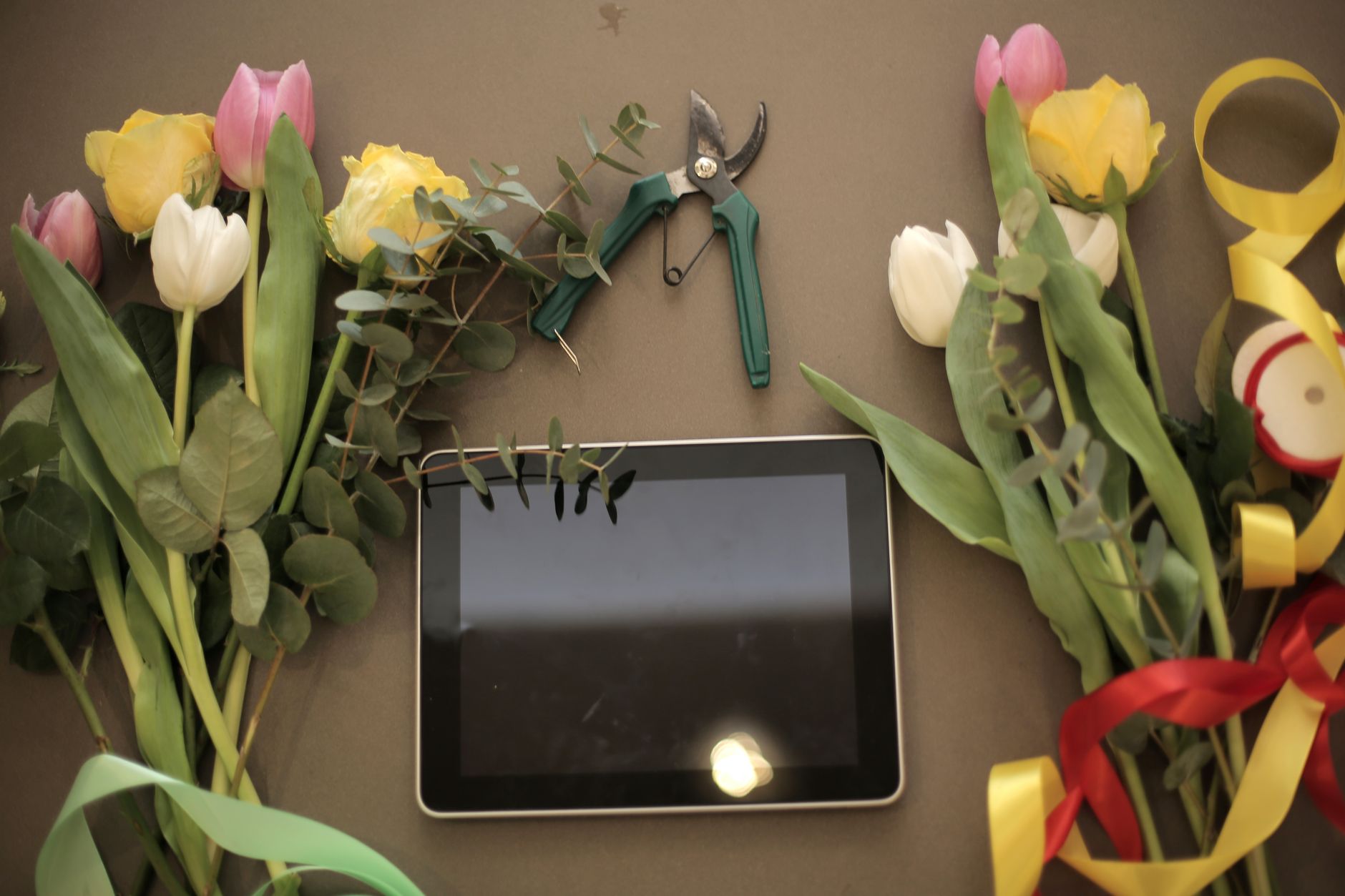 A tablet lying among tulips, roses, pruning shears, and ribbon on a gray table