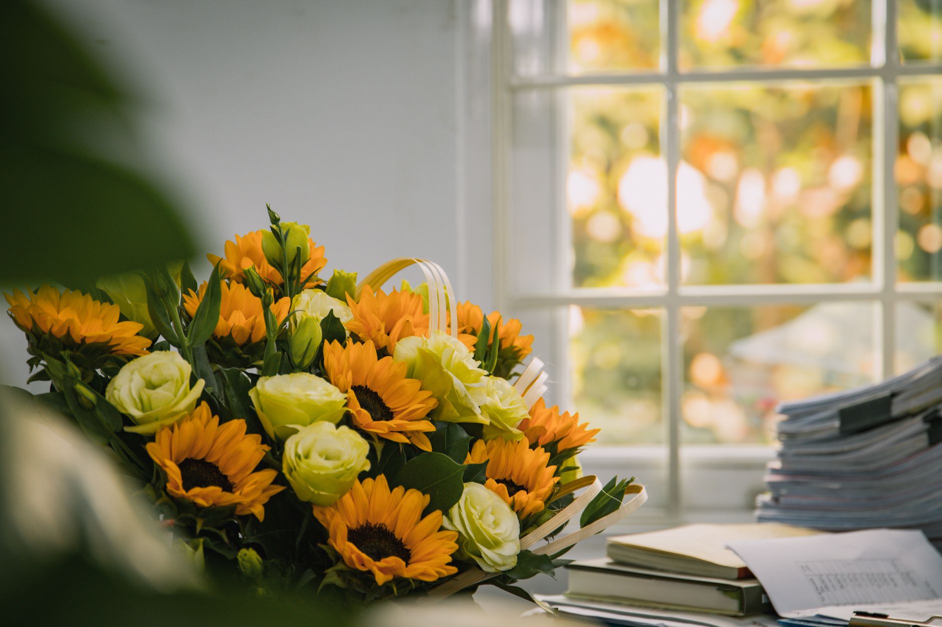 A bright bouquet of sunflowers and cream roses resting on a sunlit desk beside a stack of books